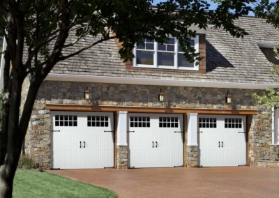 Three white residential garage doors with windows and black hardware are featured on a stone house, for installation